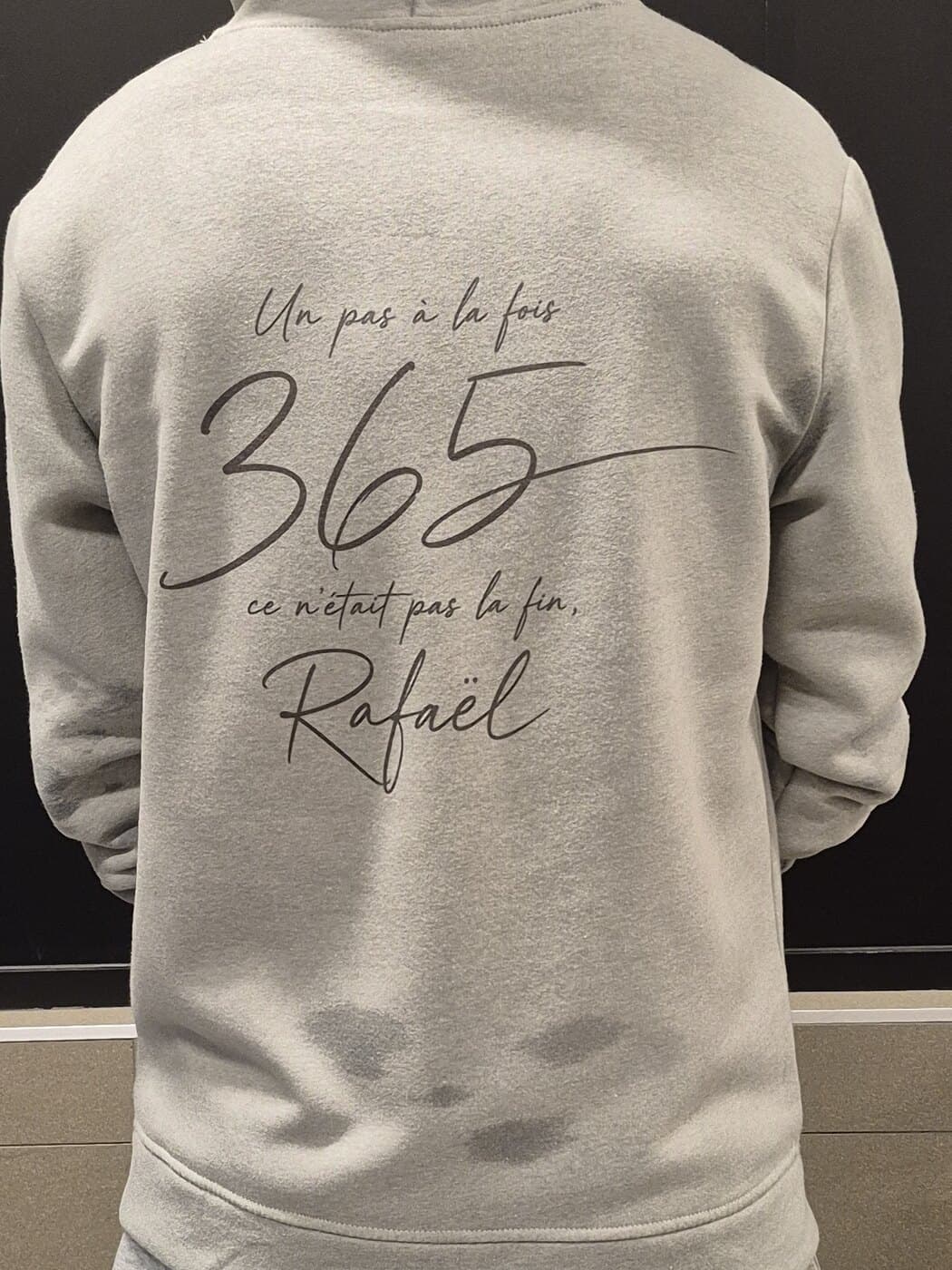The back of the grey hoodie, worn. Printed on it in a handwritten script: Un pas à la fois, then a large 365, then ce n'était pas la fin, Rafaël.
