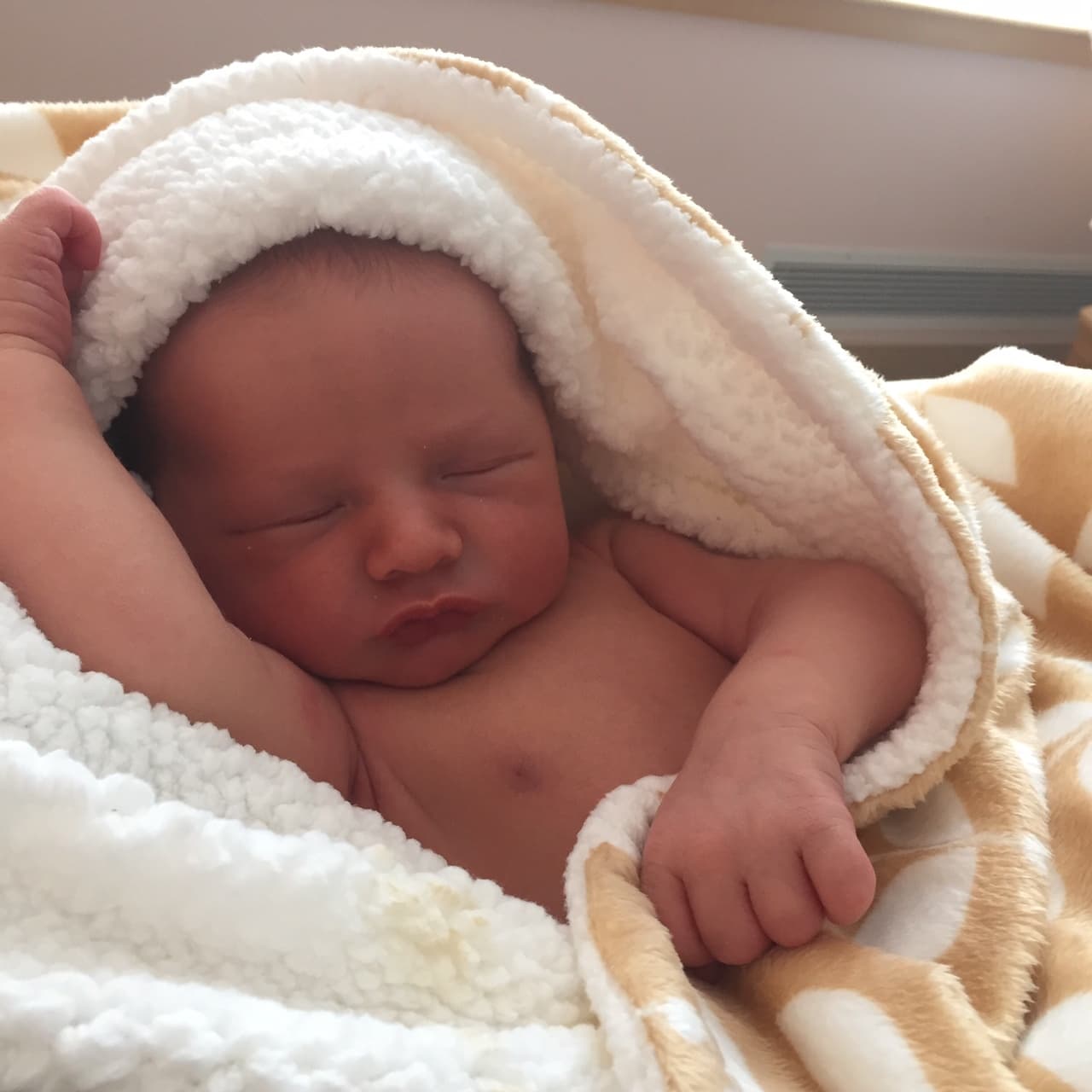 Rafaël a few days old, asleep in a white fleece blanket, one small fist up beside his head.