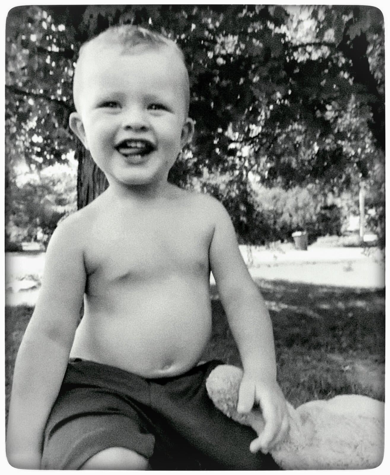 Rafaël sitting in the grass under a tree, shirtless and laughing, one hand resting on his teddy bear — the black-and-white print Dominic made.